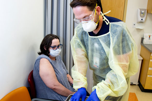 Male nurse in glove, mask, goggles, and gown preparing vaccine in doctor's office for sitting female patient
