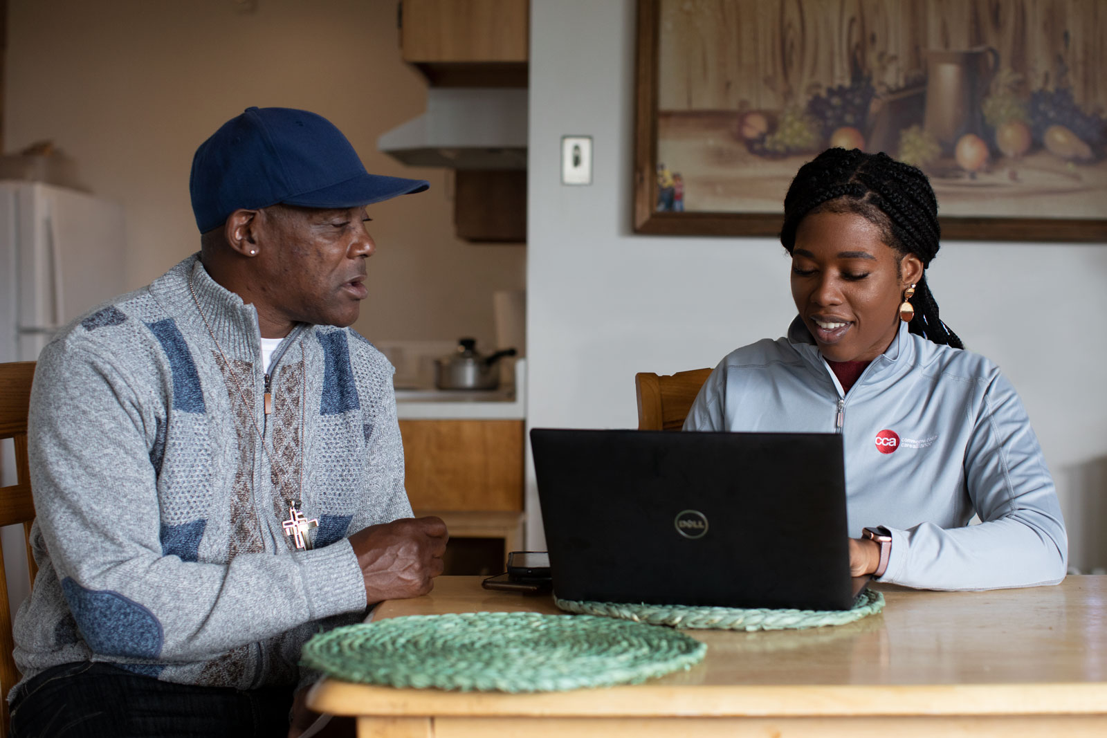 Female care partner with laptop and male member with blue baseball cap sitting at his home kitchen table