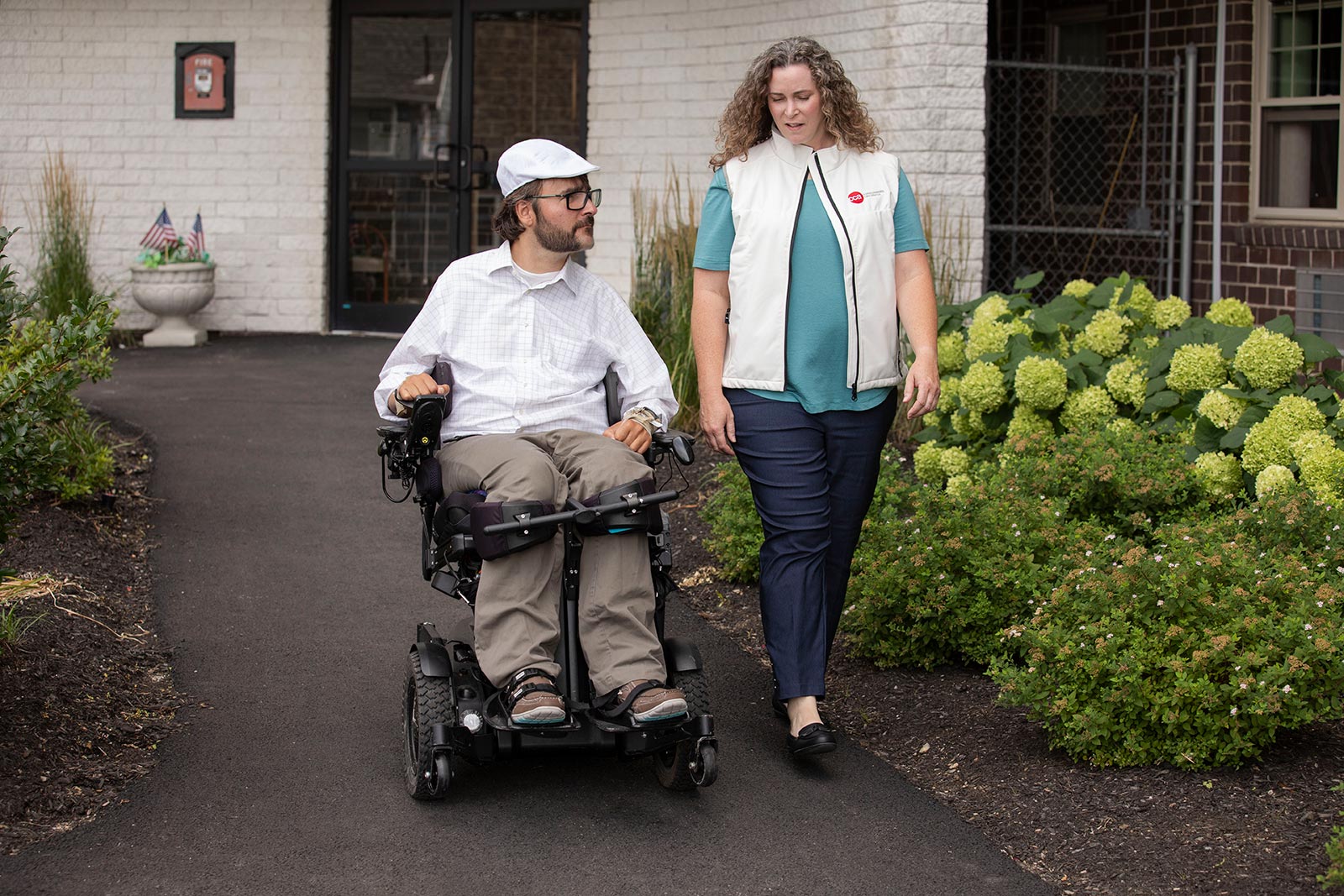 Disabled man in wheelchair and female care partner talking outdoors on walkway