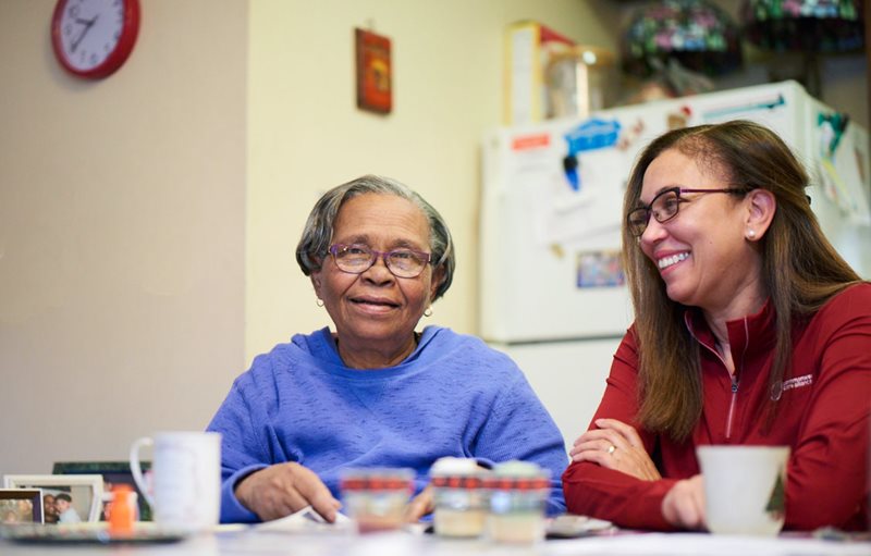 Older female member and female care provider sitting and smiling at kitchen table with coffee