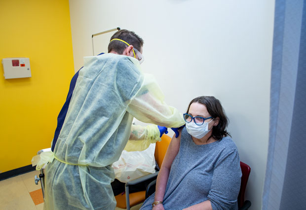 Enfermero masculino con guantes, mascarilla, antiparras y bata administrando una vacuna en el consultorio a una paciente.