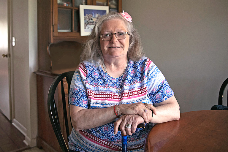 Mujer adulto mayor con los brazos cruzados sobre su bastón sonriendo en el comedor de su casa.