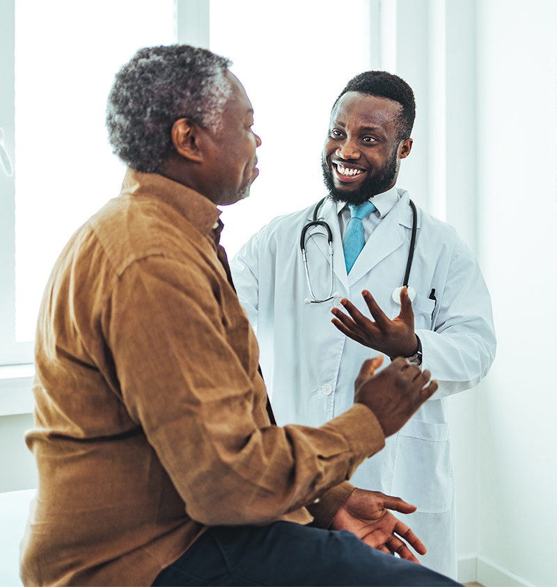 Older male patient seated with mask on listening to female nurse with mask with her hand on his shoulder at doctor's office