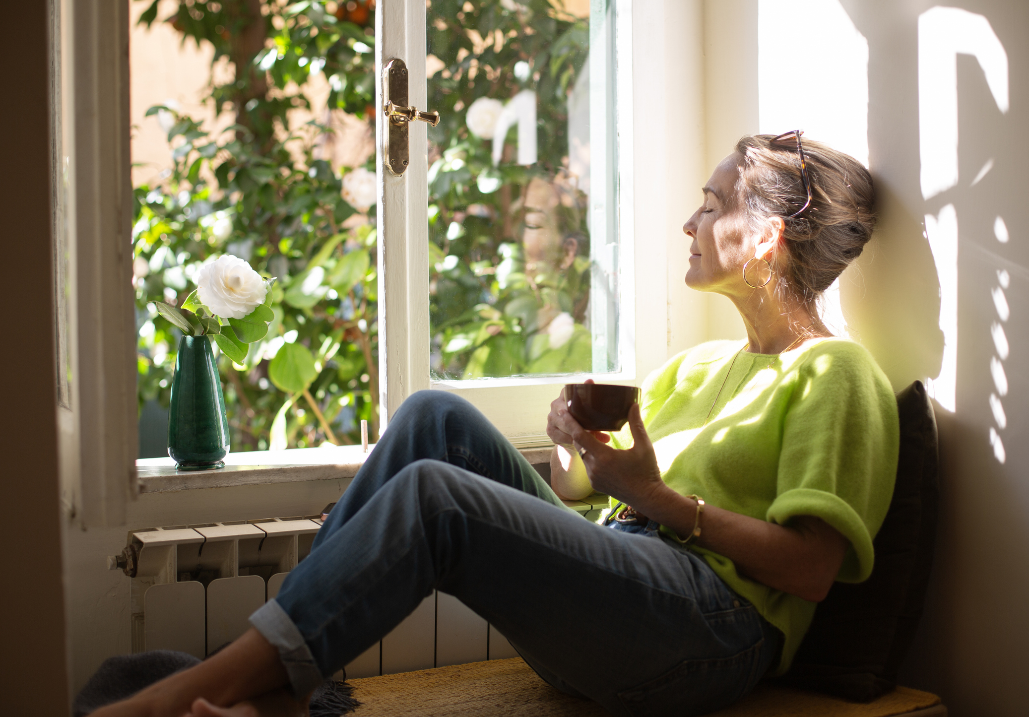 Woman relaxing in a nook by the open window, enjoying the sun and a cup of coffee