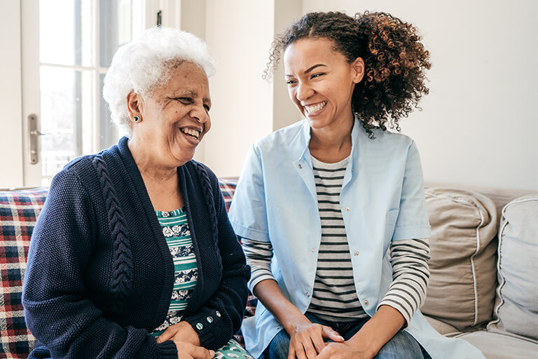 Elderly woman smiling on her couch at home with young female care provider