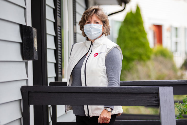Female care provider with mask in CCA vest standing on front porch of a home