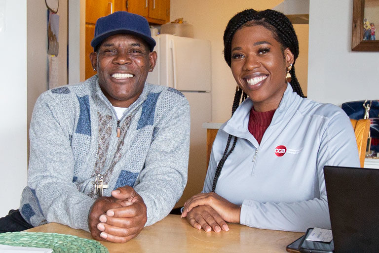 Female care provider sitting and smiling at kitchen table with middle-aged male member