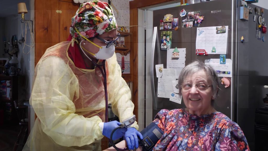 Nurse wearing PPE equipment taking blood pressure of elderly woman in her kitchen