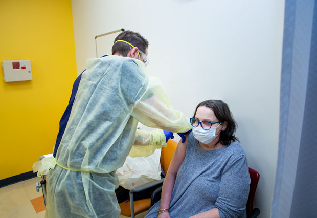 Male nurse in glove, mask, goggles, and gown administering vaccine in doctor's office for sitting female patient