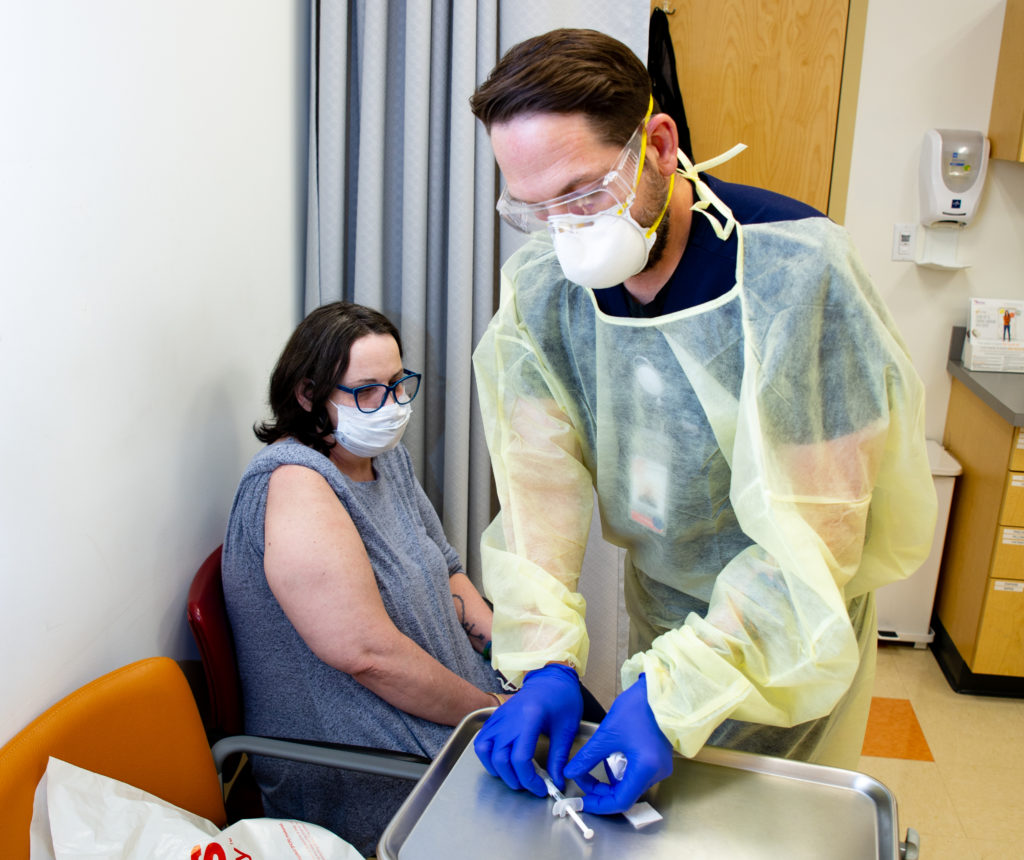 Male nurse in PPE equipment preparing vaccine in doctor's office for sitting female patient