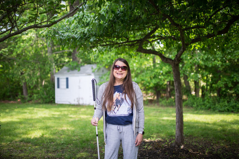 One Care member Marie standing and smiling outside with sunglasses and white cane