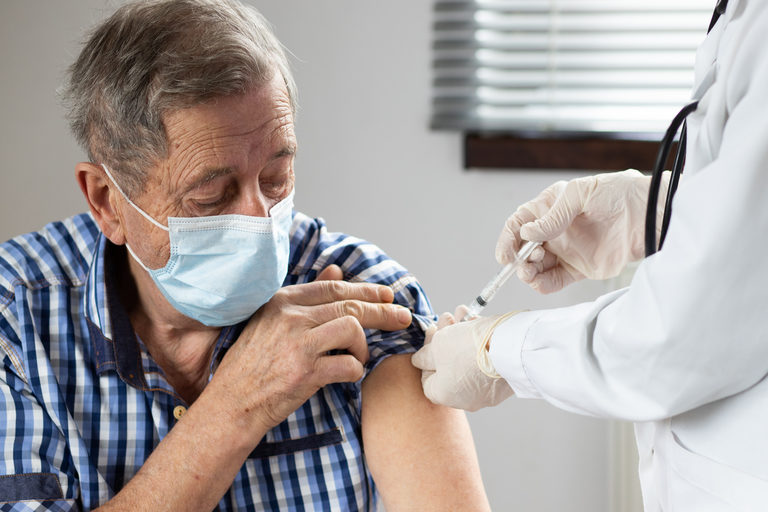 Medical worker in a white lab coat giving a shot to an older man wearing a surgical mask