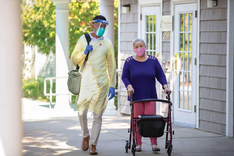 Male healthcare provider in PPE equipment walking outside with elderly female patient with mask and walker