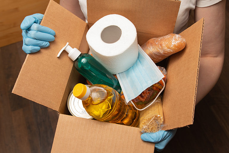Person holding cardboard box filled with a roll of toilet paper, a bottle of soap, a disposable mask, and other supplies.