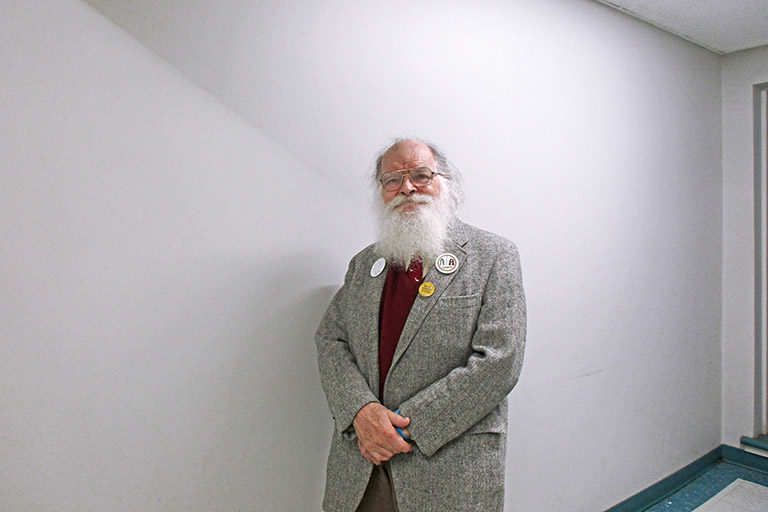 Older man with a long, white beard wearing a sports coat stands in a hallway