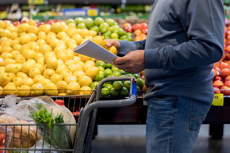 Man standing in a grocery store produce aisle with a shopping cart and grocery list