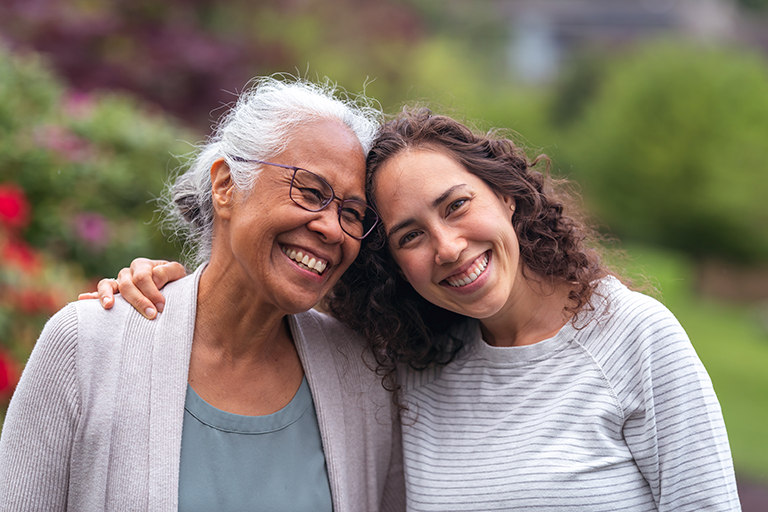 A mother and daughter standing together, hugging and smiling outside in a park.