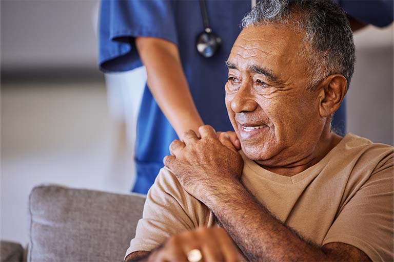 Black man wearing a tan shirt, sitting in a gray chair, smiling. Black woman wearing blue scrubs holding his hand on his shoulder.
