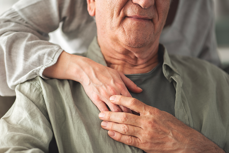 Older adult man sitting while a caregiver stands above him with her hand on his chest.